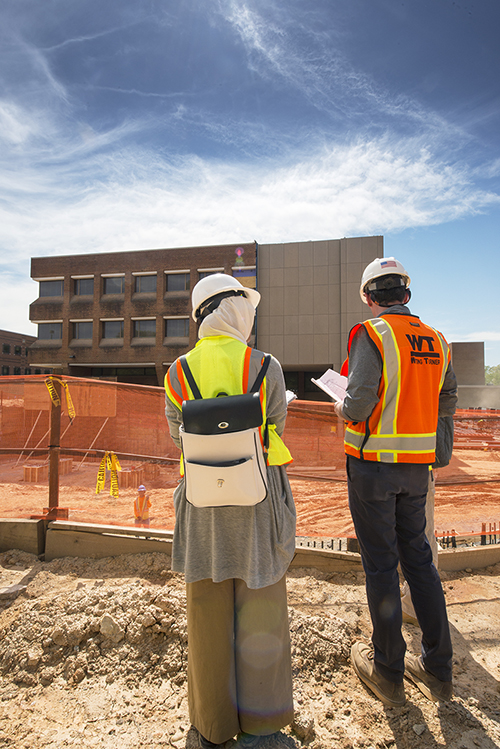 Two people standing, back to the camera, with hard hats on, looking at a construction site.  