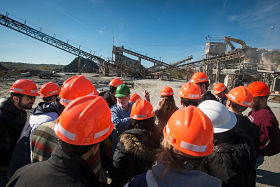 A group of students wearing orange hard hats surround their professor to listen to her instructions in a landfill