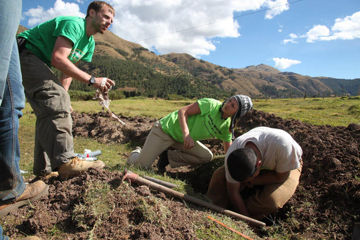 Three students digging a hole in a big open field with mountains in the background. 