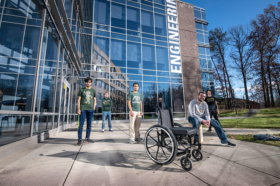 Six teammates of the senior design team stand outside in front of the engineering building with the wheelchair they are building.
