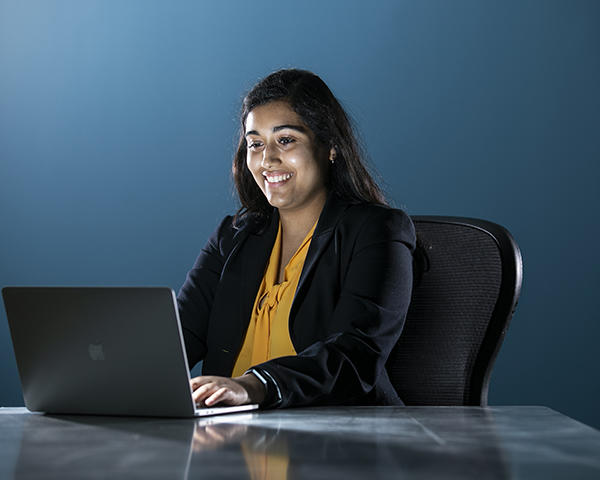 Maya Chatterjee sitting at a desk in a dark blue room with an Apple laptop in front of her.