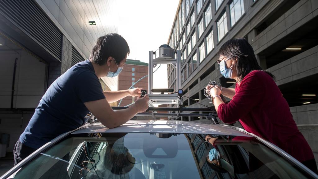 A two person team works on a piece of technology attached to the roof of a car.