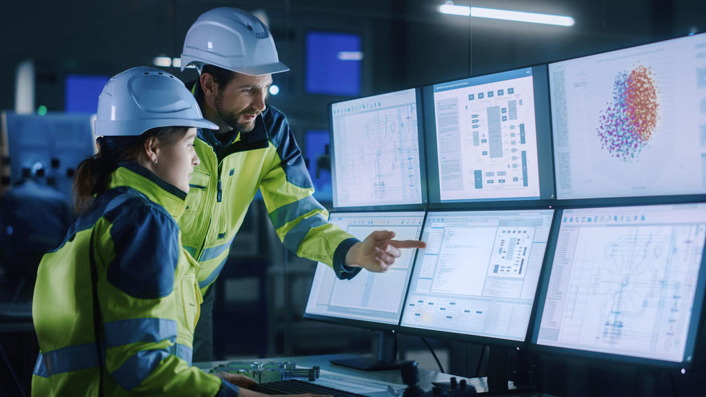 Systems Engineers wearing hard hats and yellow vests look at data points of a land project on multiple computer screens