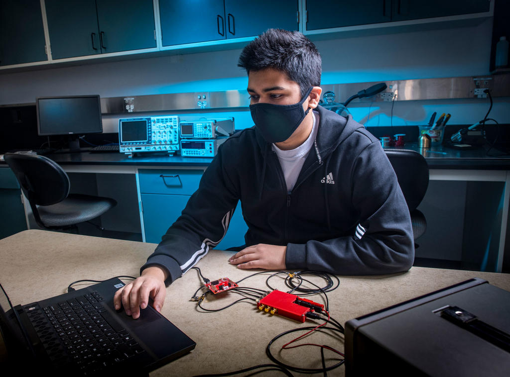South Asian male student at George Mason University wears a mask and operates a signal processing board and computer