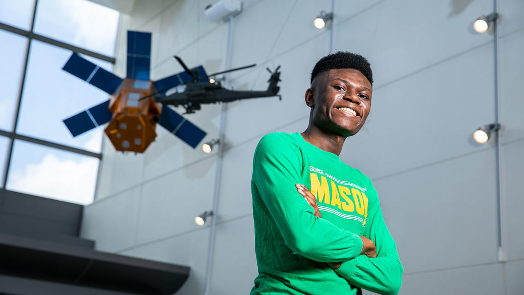 Black male student at George Mason University wears a long-sleeve shirt and smiles in front of a satellite in the background