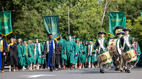 Graduates and faculty marshals march down the street under college banners, led by three drummers in colonial garb and the Mason Patriot mascot.