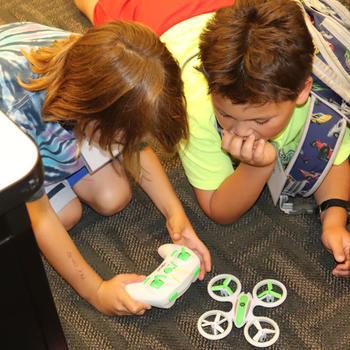 Two young boys play with a game controller to operate a miniature drone. 